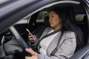 Woman in gray blazer seated in car, focused on smartphone while driving in urban Alabama—highlighting distracted driving risks and legal implications.
