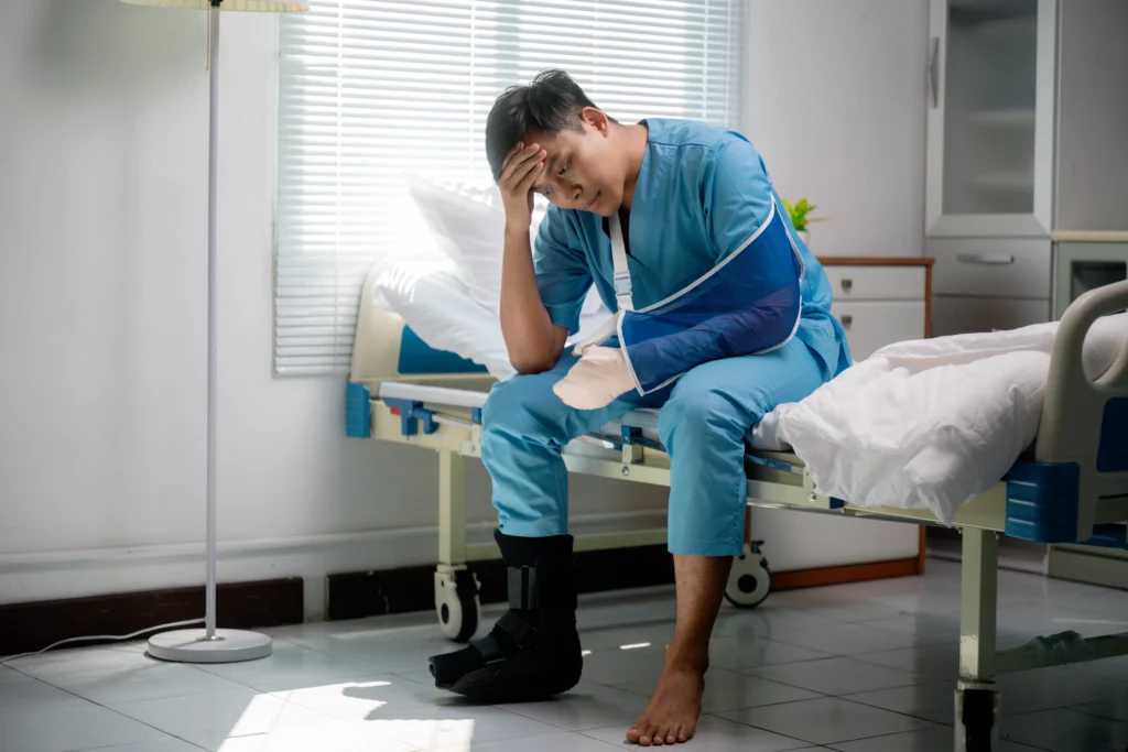 Pensive man in a hospital room with arm in a sling and boot cast, illustrating dealing with car accident medical bills in Alabama before settlement.