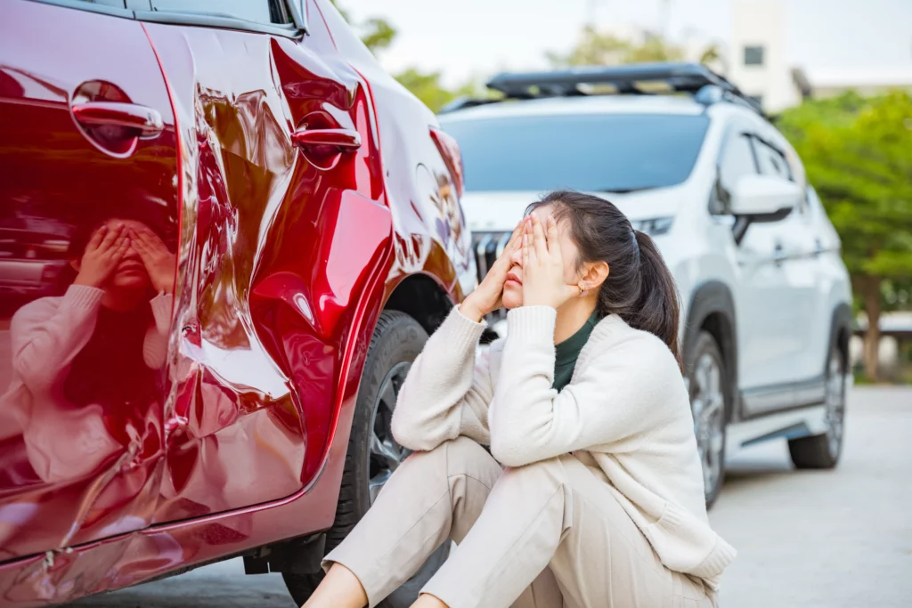 Distressed woman sitting on the ground beside a dented red car after a crash, illustrating the importance of seeing a doctor soon after a car accident in Alabama.