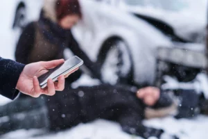 Person holding a smartphone at a snowy crash scene while another helps an injured driver near a damaged car, illustrating giving a recorded statement to insurance after an accident.